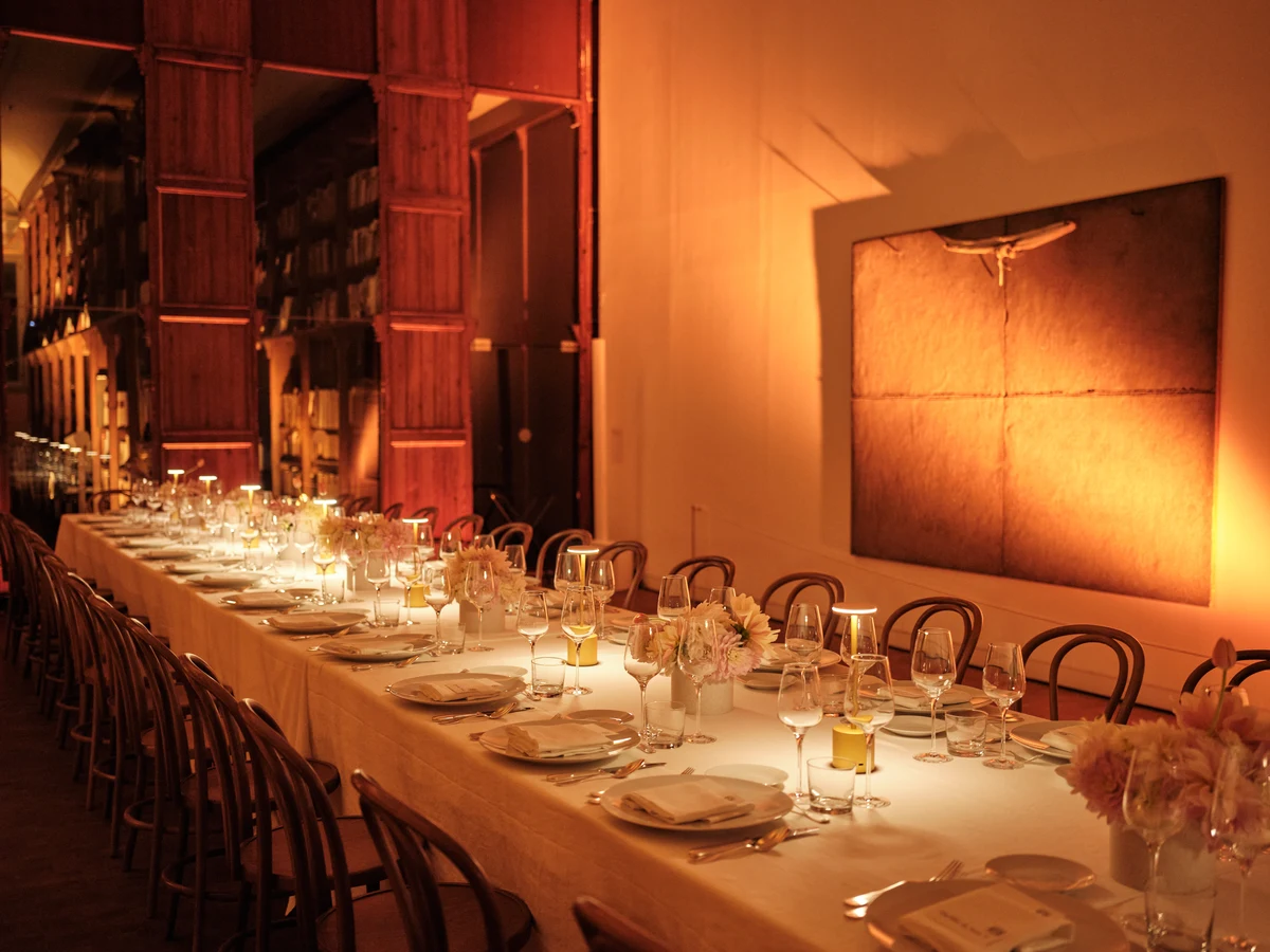 Mesa corrida de banquete con velas y copas, junto a la estantería de madera iluminada en cálido.